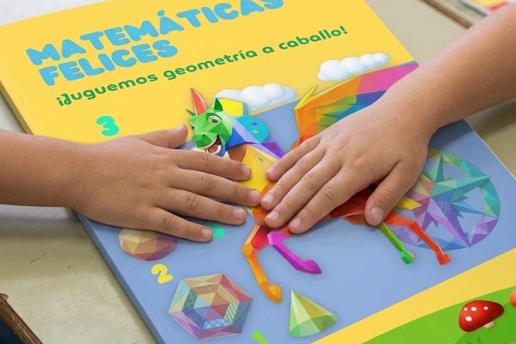 Las manos de dos niños sobre la portada de la cartilla Matemáticas Felices, en un aula